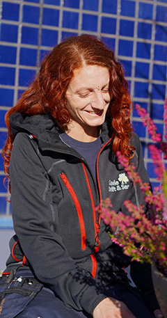 Kvinna med rött hår och mörk jacka, sitter vid plantering av lju, framför blå vägg.